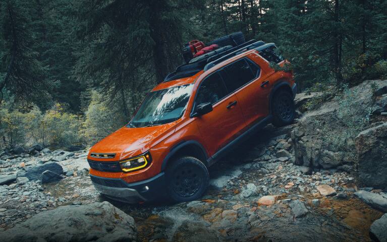 2027 Honda Passport TrailSport Elite in Sunset Orange profile on a rocky trail in a dense forest (Mid-Size SUV).